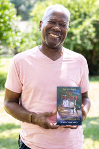 Photo of David smiling in a soft pink shirt and holding a copy of his book in a park setting.