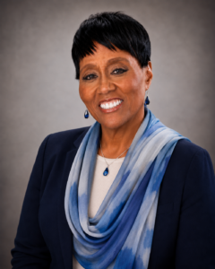 Photo of Carla Jordan smiling in a navy blazer with a blue scarf and matching jewelry against soft gray background.
